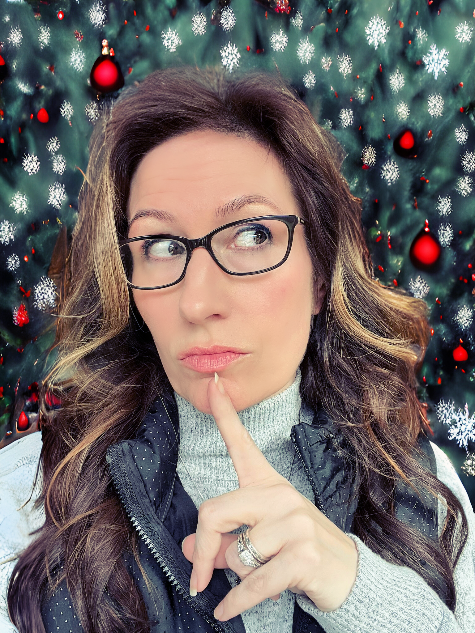 woman in glasses with finger pressed to lips, looking off camera in thoughtful expression in front of christmas tree and ornament style backdrop